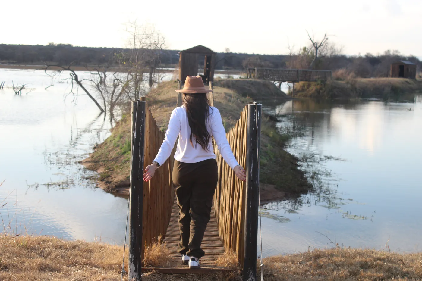 A person in a sun hat walking along a narrow wooden walkway over the water to a bird hide