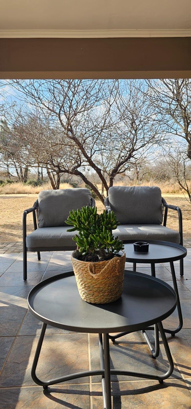 Two grey patio chairs and round tables on the covered stoep, looking out at the bush